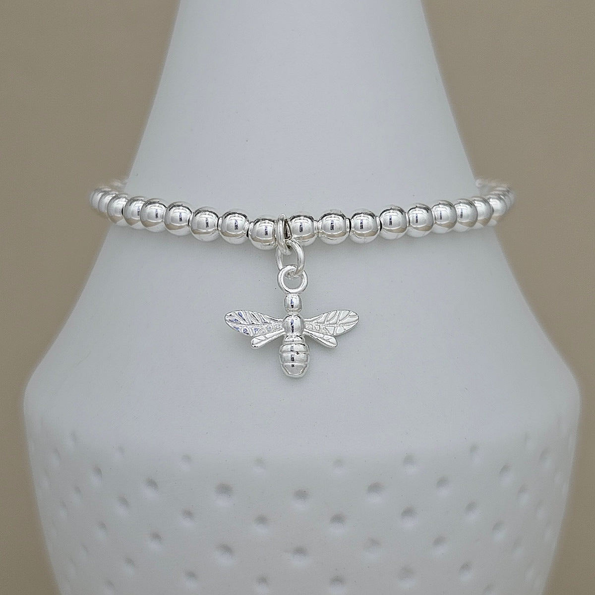 A silver beaded bracelet with a dangling bee charm, displayed on a white textured cone stand against a neutral beige background.