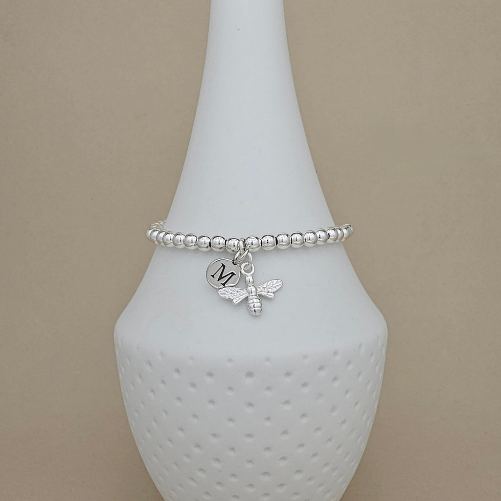 A silver beaded bracelet with a bee charm and a heart charm engraved with the letter 'M', displayed on a white textured cone stand against a beige background.