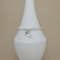 A silver beaded bracelet with a bee charm and a heart charm engraved with the letter 'M', displayed on a white textured cone stand against a beige background.