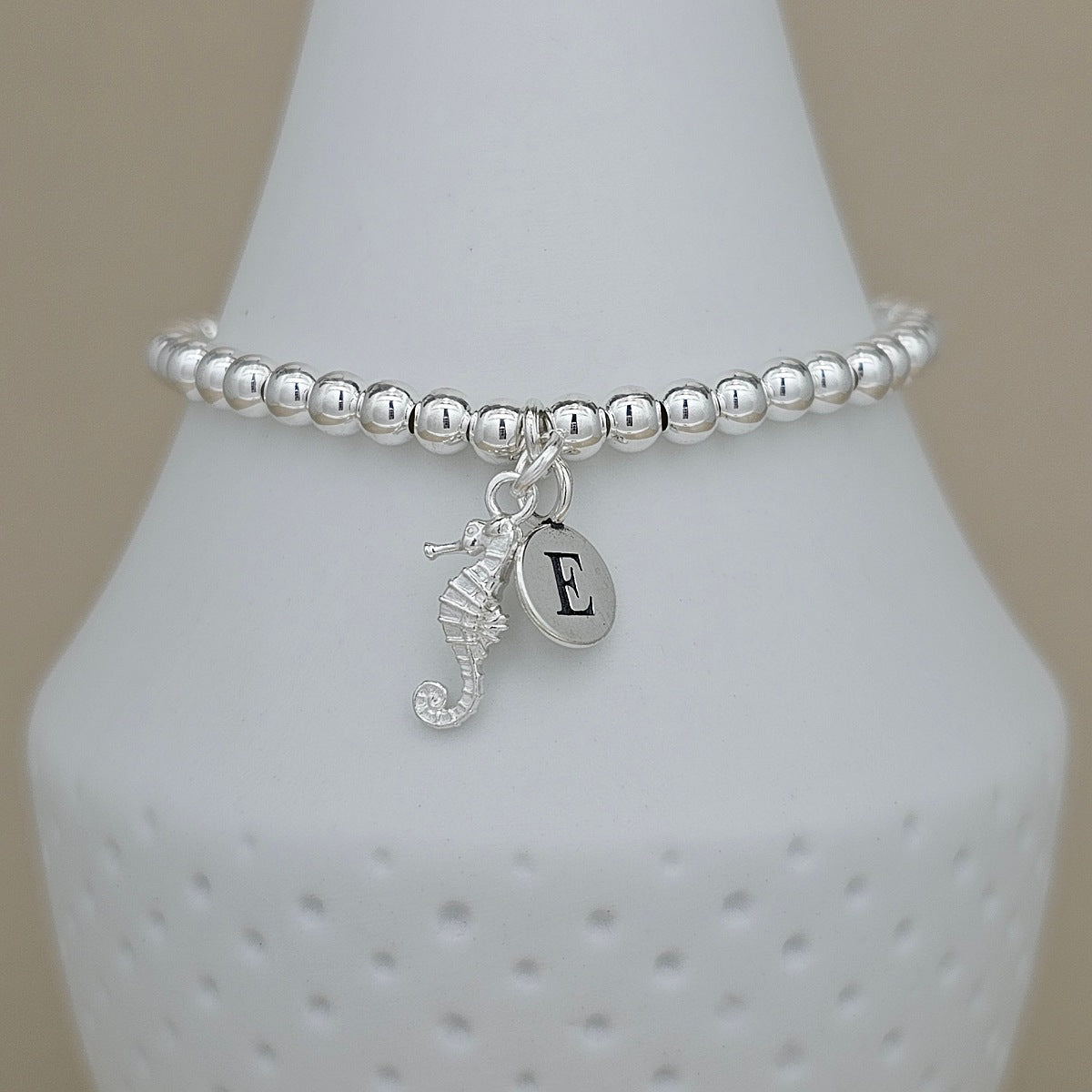A silver beaded bracelet with a seahorse charm and an initial 'E' monogram disc charm, displayed on a white ceramic jewelry cone against a beige background.
