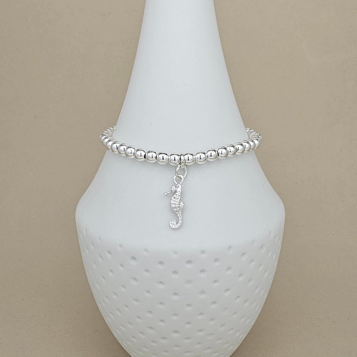 A silver beaded bracelet with a dangling seahorse charm, displayed on a white ceramic jewelry cone against a beige background.