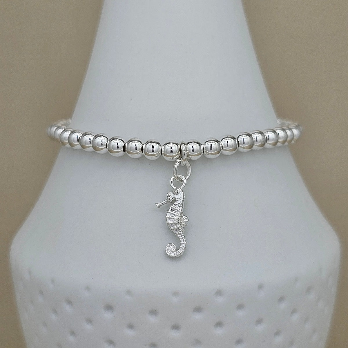 A silver beaded bracelet with a small seahorse charm, displayed on a white cone-shaped jewelry stand against a warm beige background.