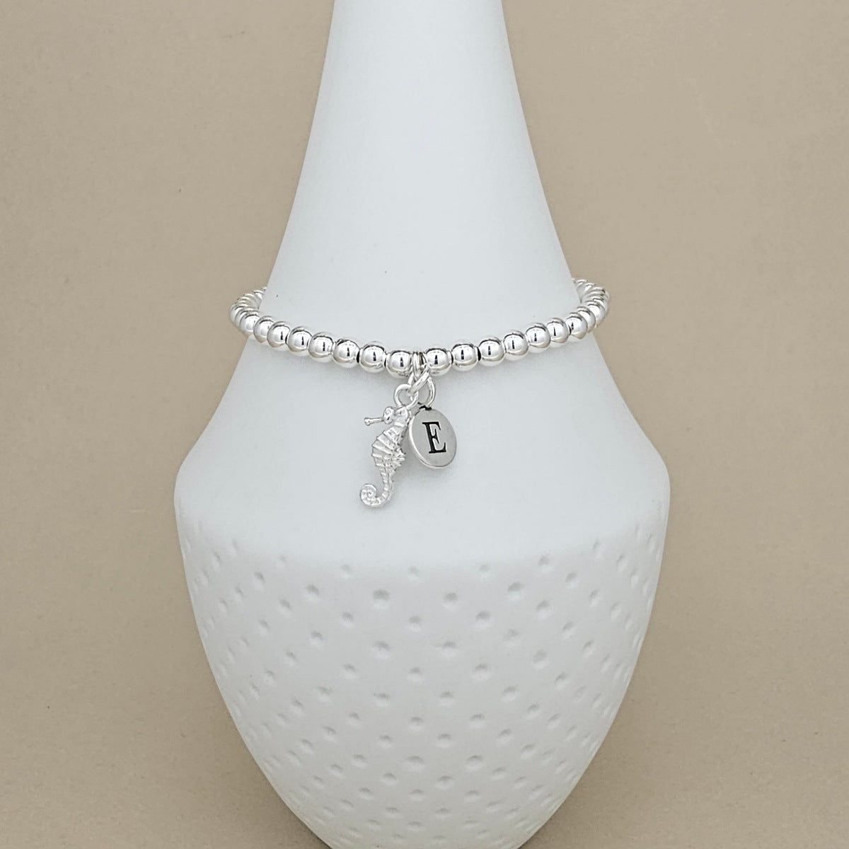 A silver beaded bracelet with a seahorse charm and an initial 'E' charm, displayed on a white textured cone stand against a beige background.