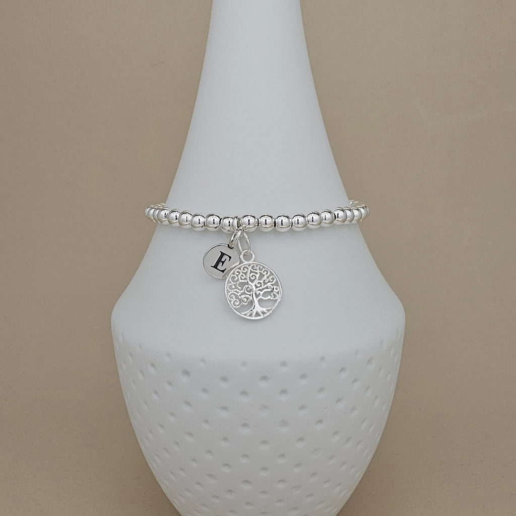 A silver beaded bracelet with a tree of life charm and an initial 'E' charm, displayed on a white ceramic vase against a beige background.