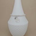 A silver beaded bracelet with a tree of life charm and an initial 'E' charm, displayed on a white ceramic vase against a beige background.