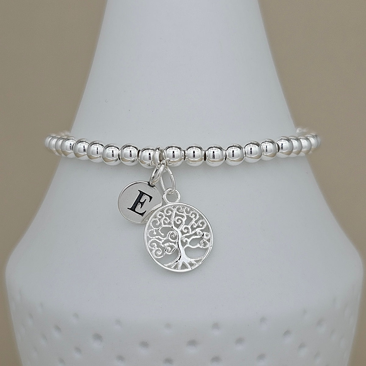 A silver beaded bracelet with a tree of life charm and an initial 'E' disc charm, displayed on a white cone stand against a beige background.