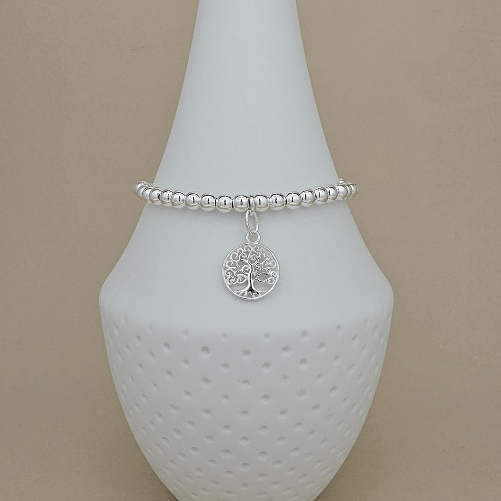A silver beaded bracelet with a circular tree of life pendant, displayed on a white ceramic jewelry stand against a beige background.
