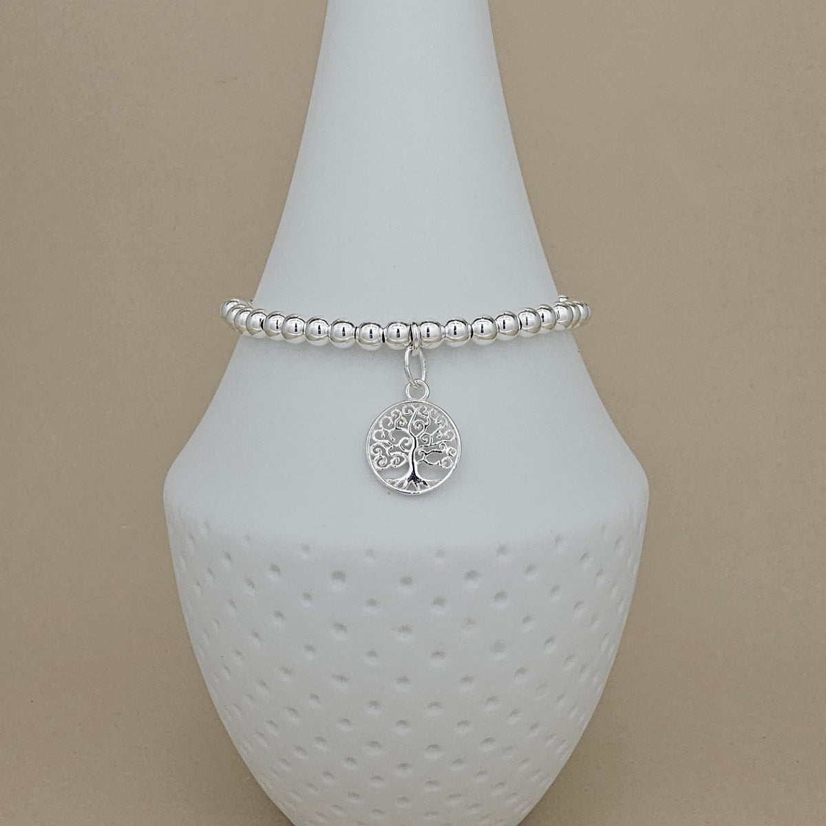 A silver beaded bracelet with a circular tree of life pendant, displayed on a white ceramic jewelry stand against a beige background.