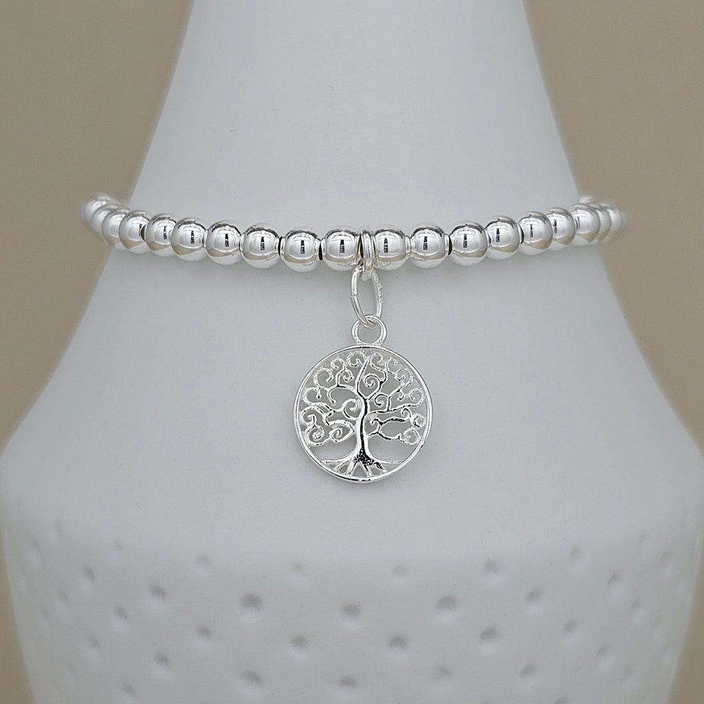 A silver beaded stretch bracelet with a circular tree of life pendant, displayed on a white ceramic jewelry cone against a neutral beige background.