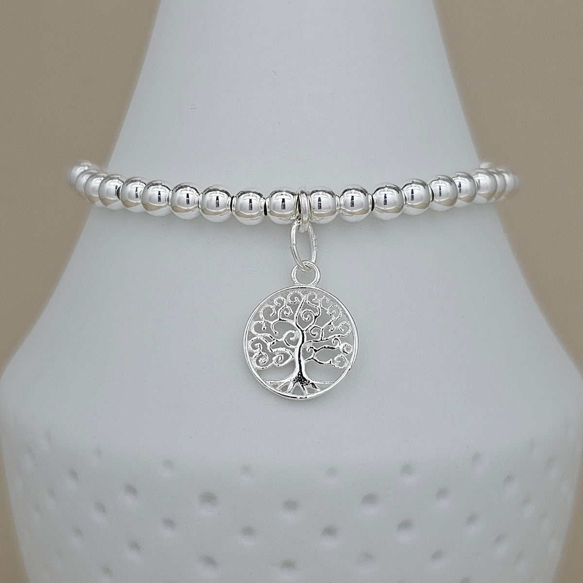 A silver beaded stretch bracelet with a circular tree of life pendant, displayed on a white ceramic jewelry cone against a neutral beige background.