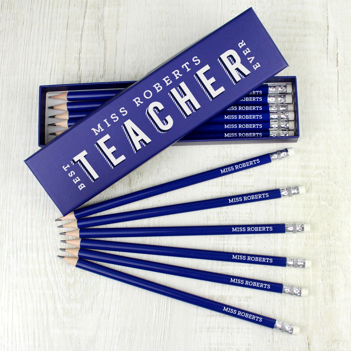 A set of personalized blue pencils with silver ferrules and white erasers, arranged in front of their matching blue packaging box. The box is labeled 'MISS ROBERTS TEACHER' in white text.