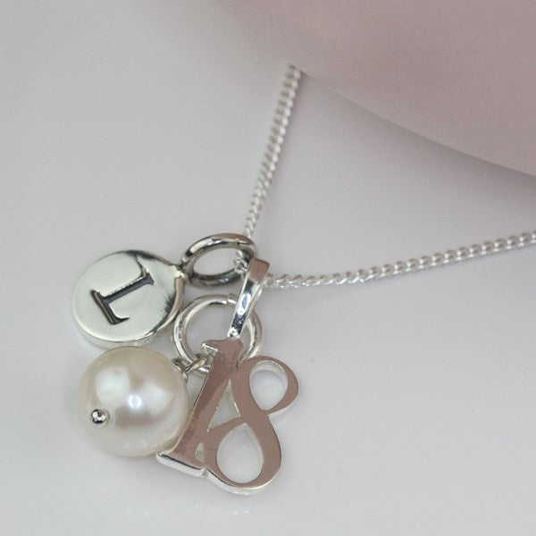 A silver chain necklace with three charms: a round silver disc engraved with the letter 'I', a white pearl bead, and a silver monogram charm reading '18'.