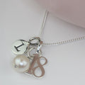 A silver chain necklace with three charms: a round silver disc engraved with the letter 'I', a white pearl bead, and a silver monogram charm reading '18'.