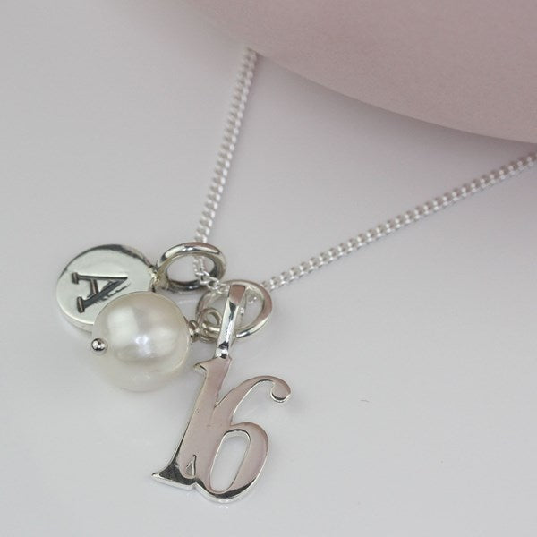 A silver chain necklace with a charm set featuring a round pearl, a silver disc engraved with the letter 'A', and a polished silver initial '16' pendant, displayed on a light gray surface.