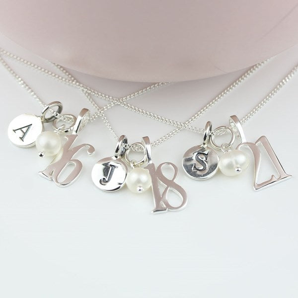 Four silver chain necklaces with personalized charms, including letters and numbers, arranged on a light pink surface.