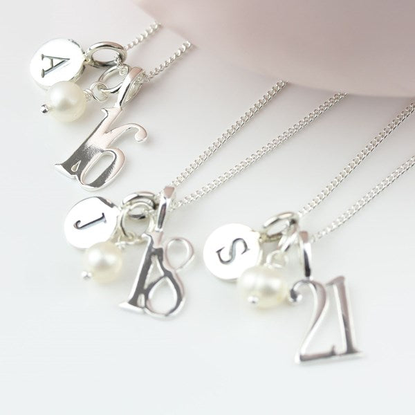 Four silver chain necklaces with personalized charms, including letters, pearls, and numerals (A, 16, J, 18, S, 21), arranged on a white surface with a soft pink curve in the background.