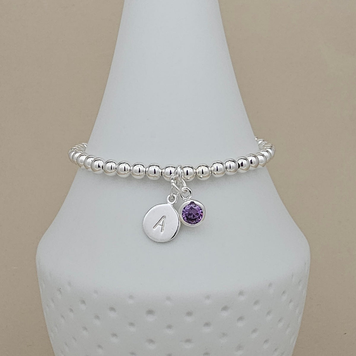 A silver beaded stretch bracelet with a round silver initial charm engraved with the letter 'A' and a small round purple gemstone charm, displayed on a white ceramic cone stand against a beige background.