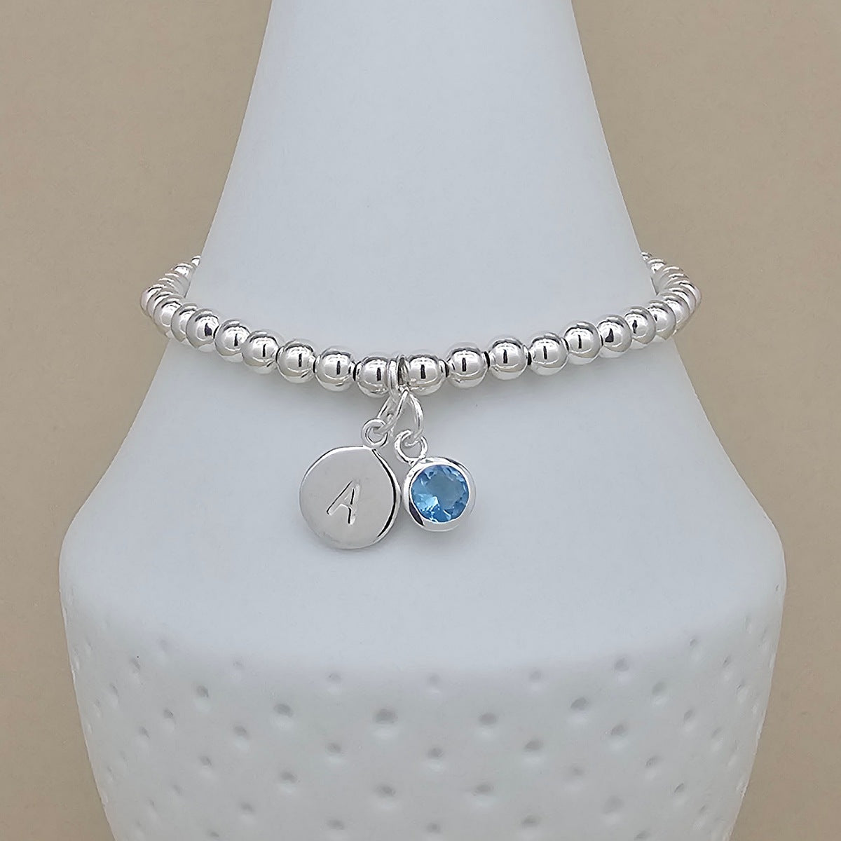 A silver beaded stretch bracelet with two charms, including a round silver disc engraved with the letter 'A' and a small round blue gemstone charm, displayed on a white cone stand against a beige background.