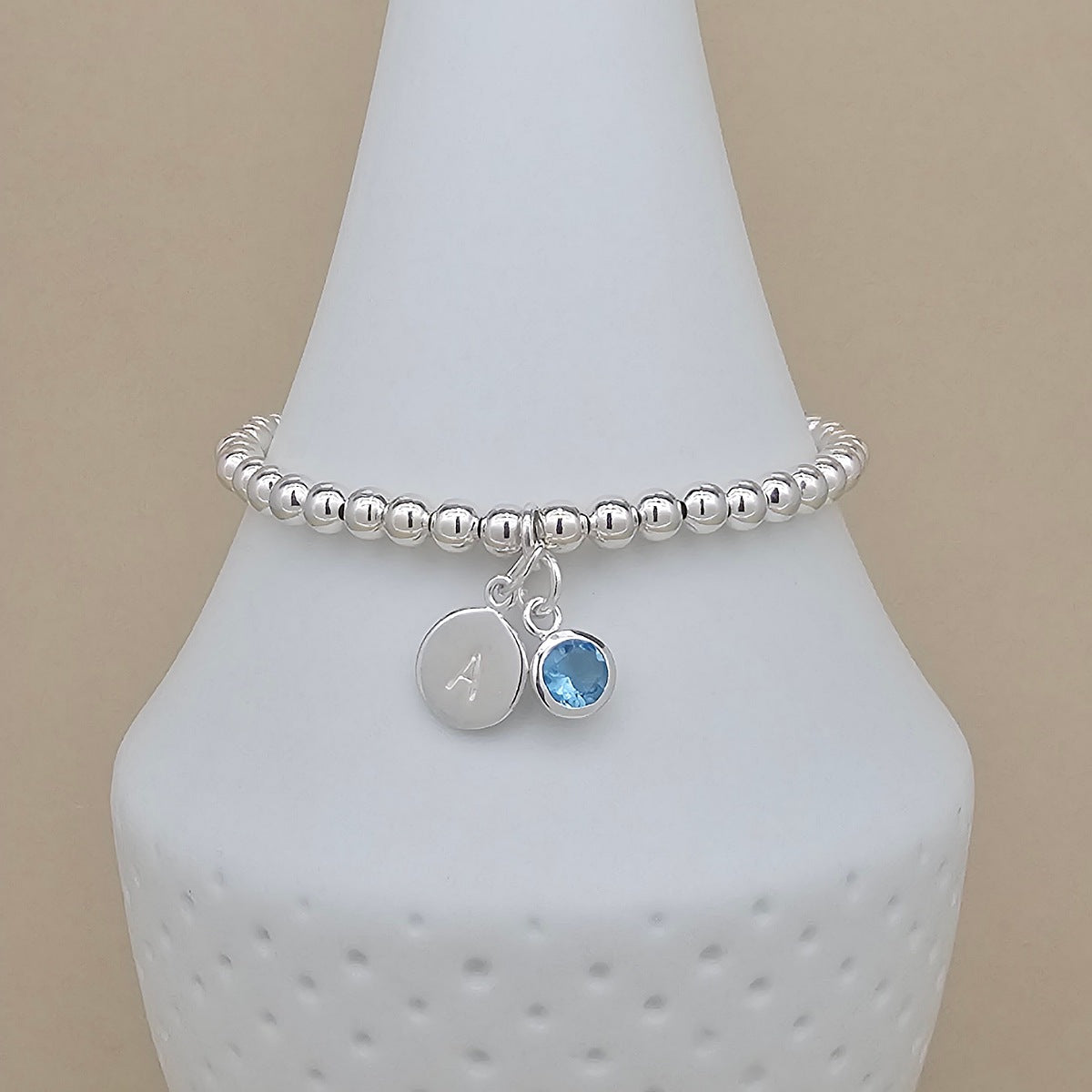 A silver beaded stretch bracelet with two charms, including a round silver disc engraved with the letter 'A' and a small round blue gemstone charm, displayed on a white cone stand against a beige background.