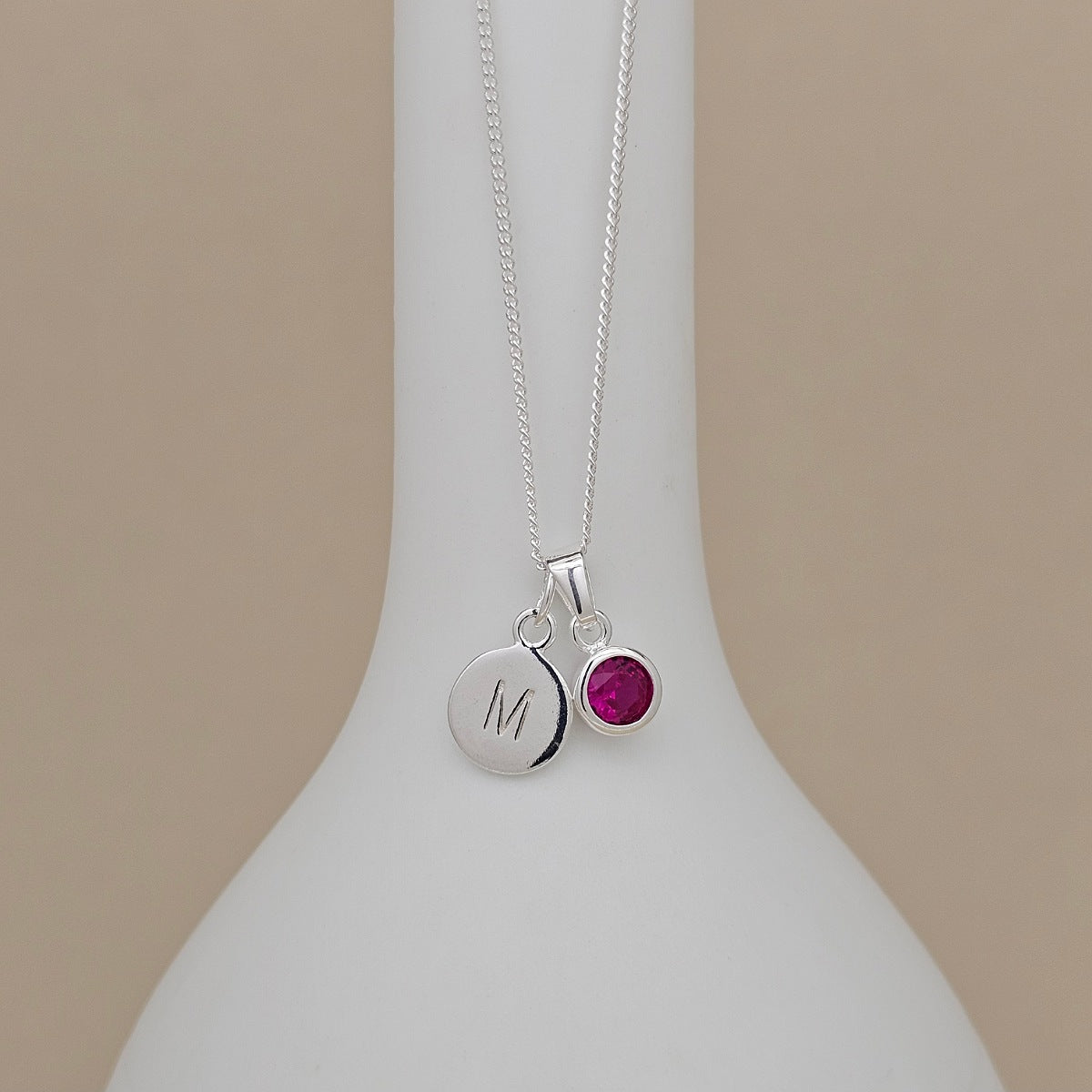 A silver necklace with two pendants displayed on a white vase: a round engraved silver disc with the letter 'M' and a small round magenta pink gemstone bezel pendant, set against a solid beige background.