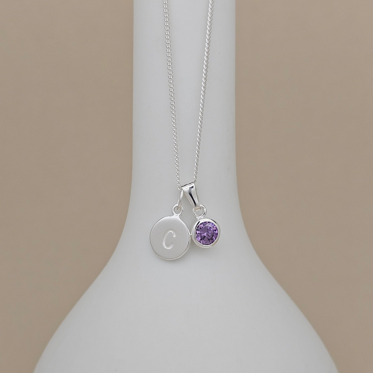 A silver necklace with a delicate chain draped over a white display bust. The pendant features two charms: a flat round silver disc engraved with the letter 'C' and a small round bezel-set purple gemstone charm.