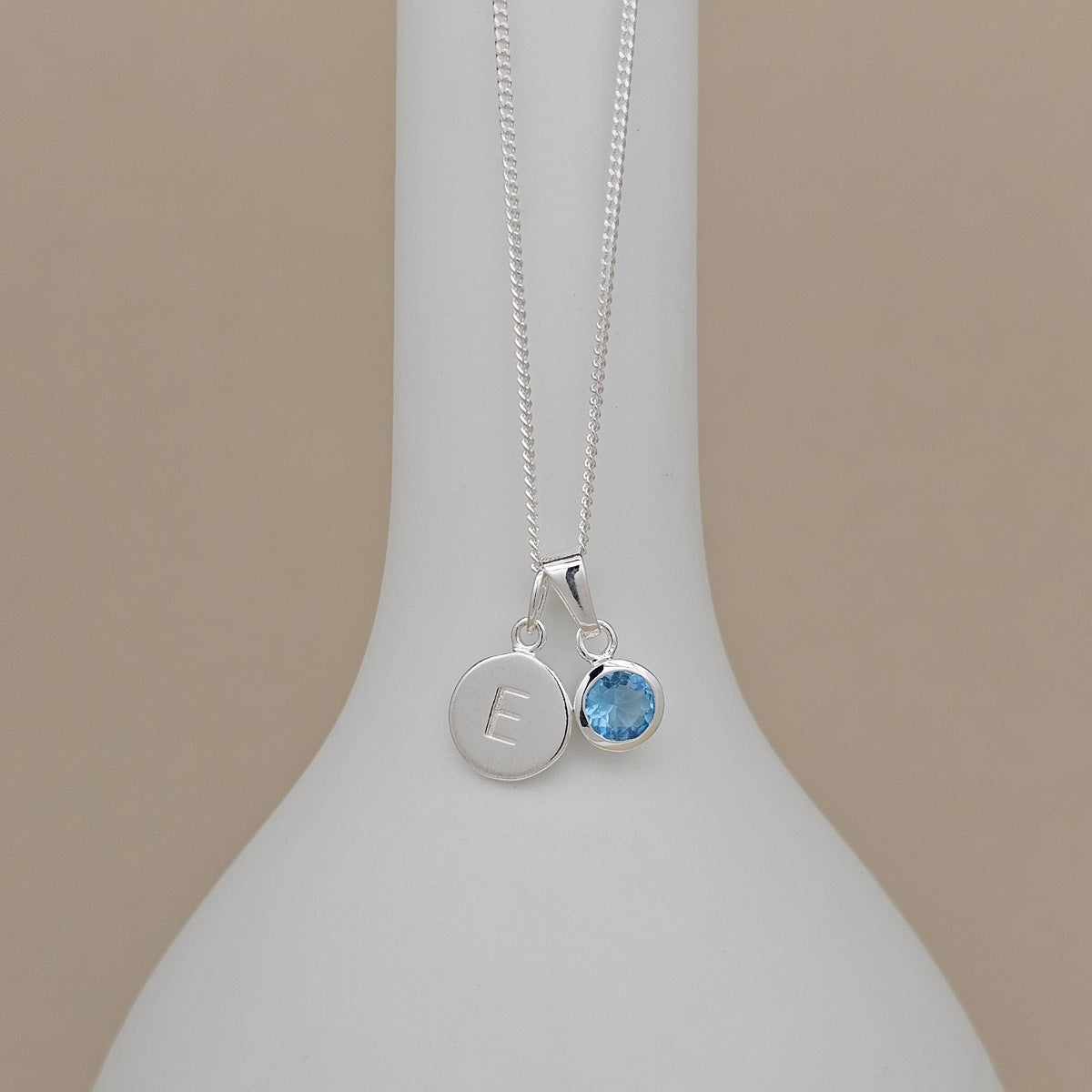 A silver necklace with a delicate chain displayed on a white ceramic stand. The pendant features a round silver disc engraved with the letter 'E' and a small bezel-set light blue gemstone.
