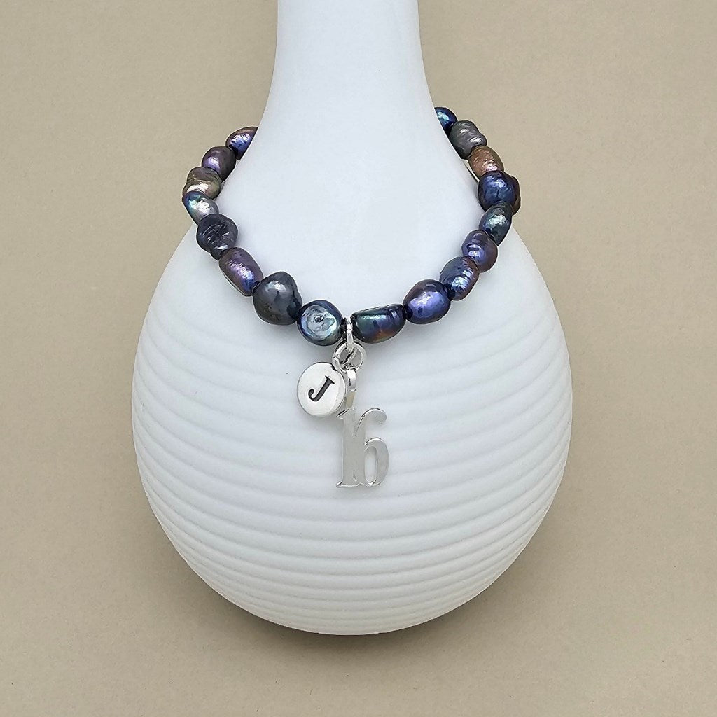 A beaded bracelet with iridescent dark blue, purple, and gray baroque-style beads, featuring a silver charm with the letter 'J' and a silver '16' initial pendant, displayed on a white ribbed vase against a beige background.