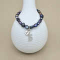 A beaded bracelet with iridescent dark blue, purple, and gray baroque-style beads, featuring a silver charm with the letter 'J' and a silver '16' initial pendant, displayed on a white ribbed vase against a beige background.