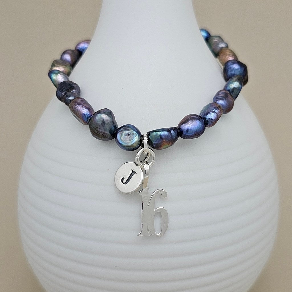 A beaded bracelet with iridescent baroque pearls in shades of blue, purple, and green, accented by a silver charm featuring the letters 'J' and '16', displayed on a white ribbed vase against a beige background.