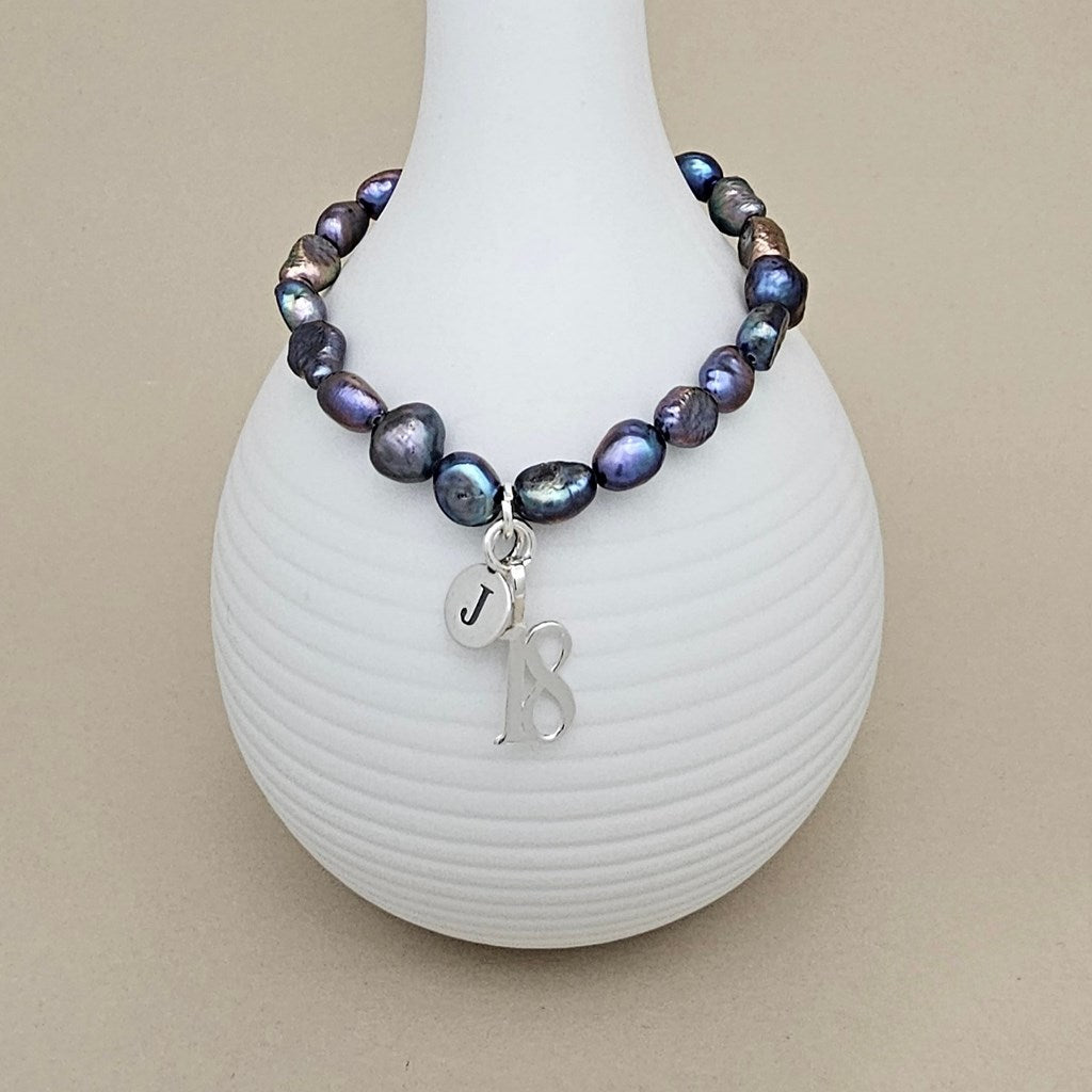 A beaded bracelet with iridescent blue, purple, and brown baroque-style beads, featuring a silver charm with the letter 'J' and '18', displayed on a white ribbed vase against a beige background.