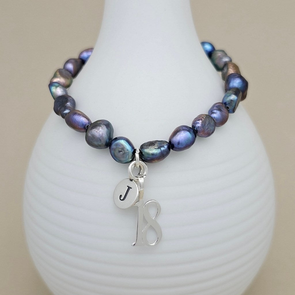 A beaded bracelet with iridescent baroque pearls in shades of blue, purple, and green, featuring a silver-tone initial 'J' charm and a '18' charm, displayed on a white ribbed vase.