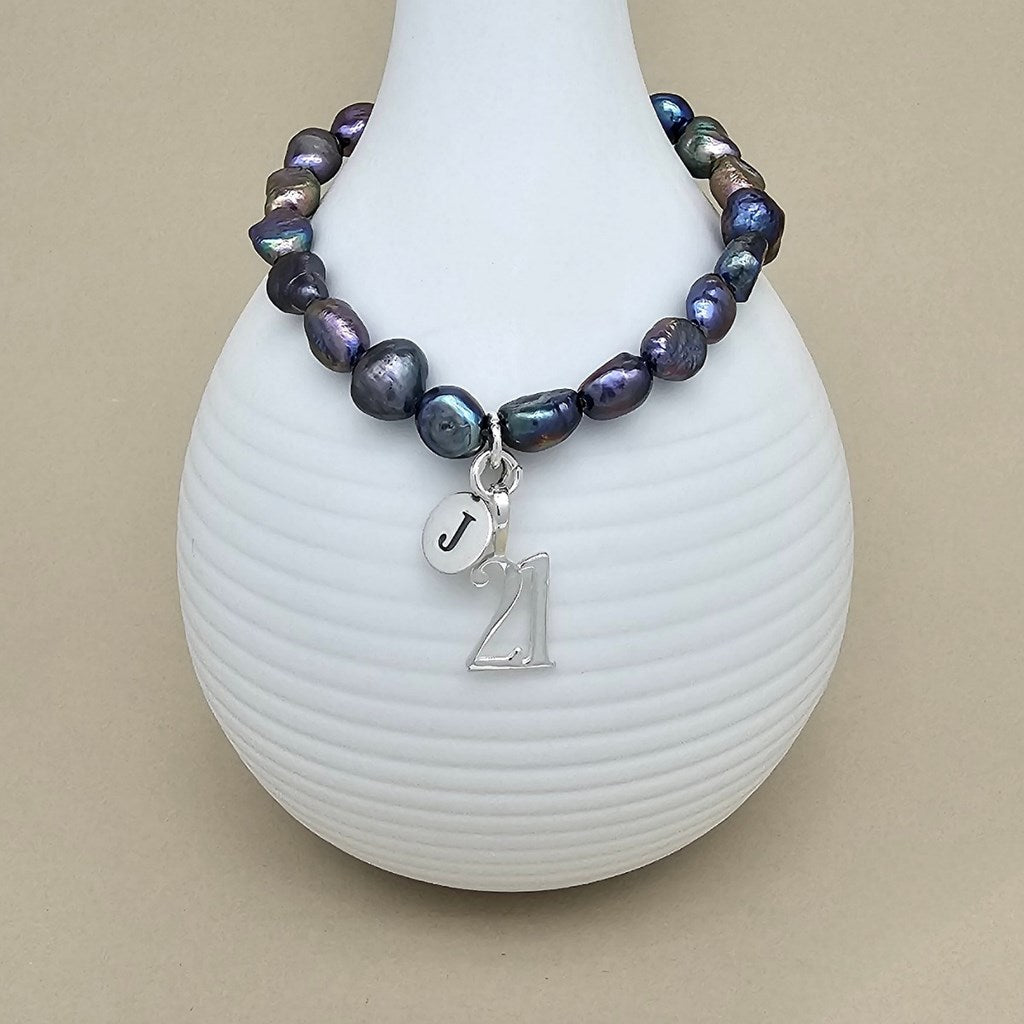 A beaded bracelet with irregular dark purple and blue baroque pearls, a silver charm with the letter 'J', and a silver '21' charm, displayed on a white ribbed vase against a beige background.