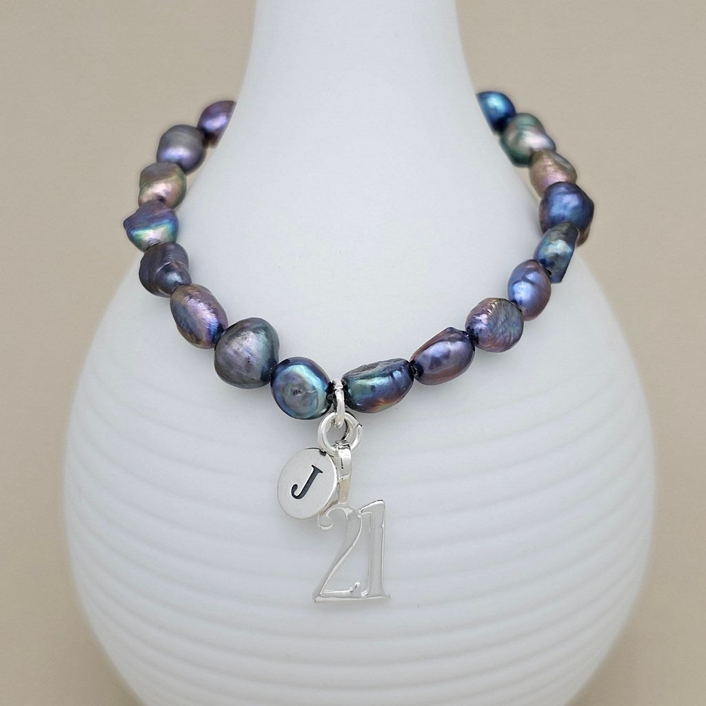 A beaded bracelet with iridescent baroque pearls in shades of blue, purple, and green, featuring a silver-tone initial 'J' charm and a '21' charm, displayed on a white ribbed ceramic vase.