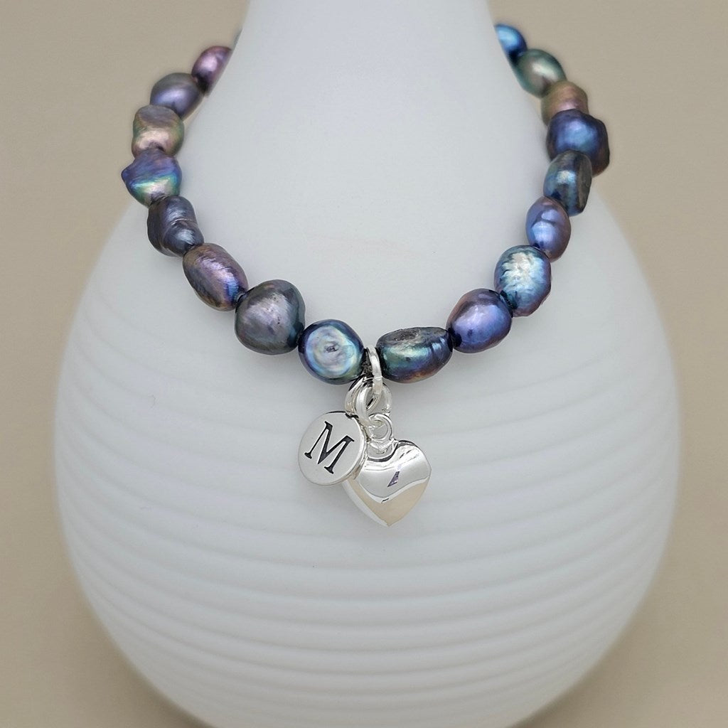A beaded bracelet with iridescent baroque pearls in shades of purple, blue, and green, featuring a silver heart charm engraved with a monogram letter 'M', displayed on a white ribbed vase against a neutral beige background.