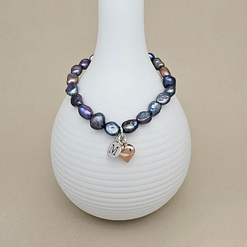A beaded bracelet with iridescent baroque pearls in shades of purple, blue, and gray, featuring a silver initial 'M' charm and a rose gold heart charm, displayed on a white ribbed vase against a beige background.