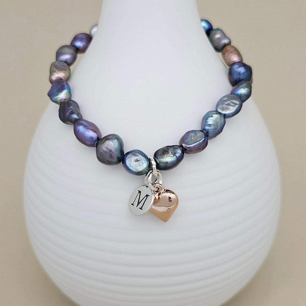 A beaded necklace with irregular baroque pearls in shades of blue, purple, and gray, featuring a silver initial 'M' charm and a rose gold heart charm, displayed on a white ribbed vase.