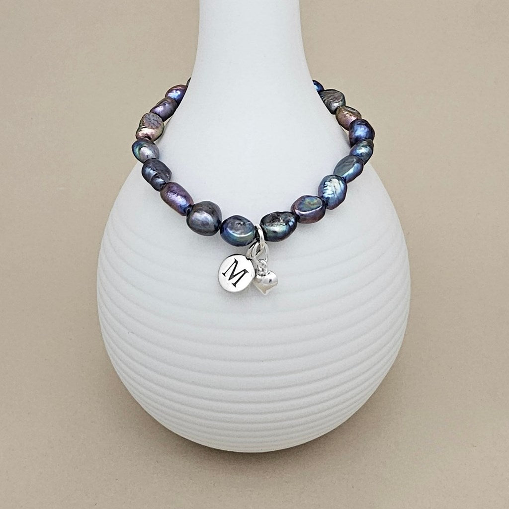 A beaded bracelet with iridescent baroque pearls in shades of blue, purple, and gray, featuring a silver charm with a monogram 'M' and a small heart, displayed on a white ribbed vase against a beige background.