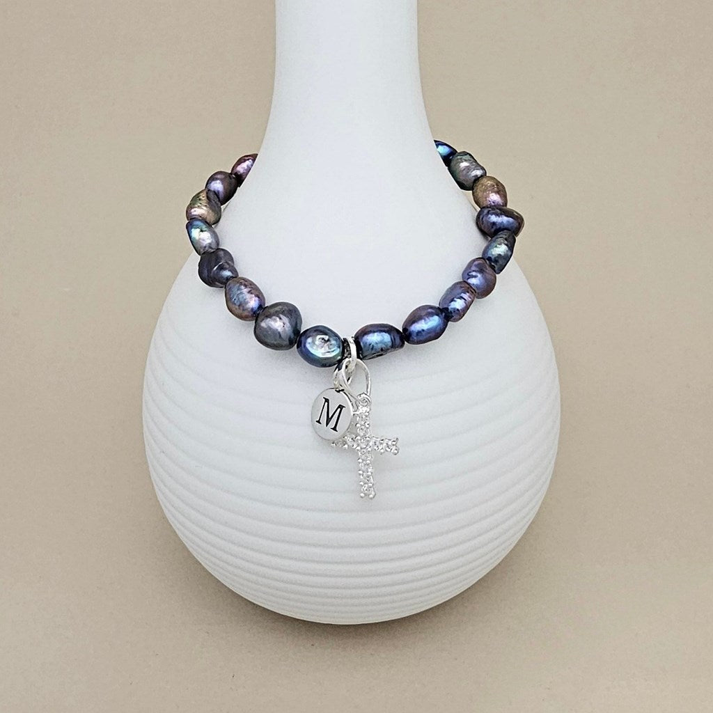 A beaded bracelet with iridescent baroque pearls in shades of blue, purple, and gray, featuring a silver-tone cross charm and a heart charm engraved with the letter 'M', displayed on a white ribbed vase against a beige background.