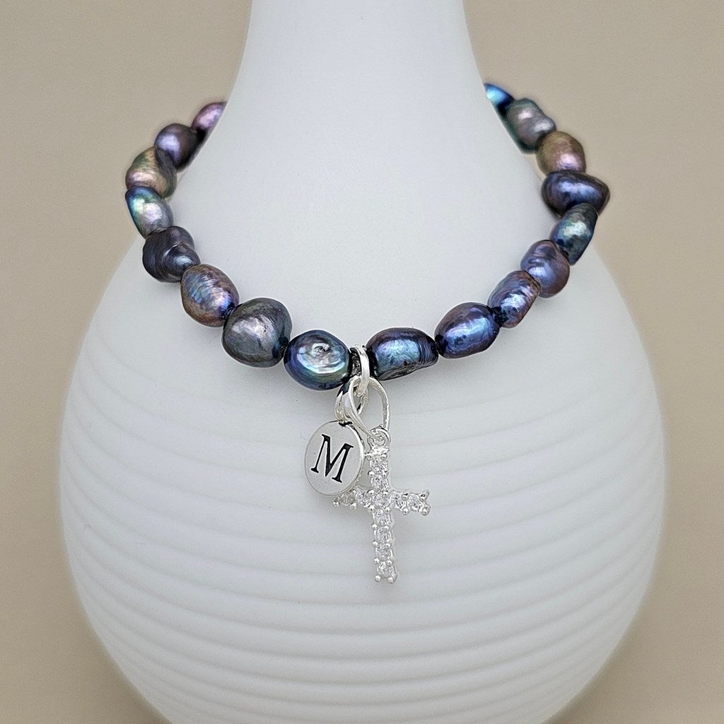 A beaded bracelet with iridescent baroque pearls in shades of blue, purple, and green, featuring a silver cross charm and a silver oval charm engraved with the letter 'M', displayed on a white ribbed vase against a beige background.