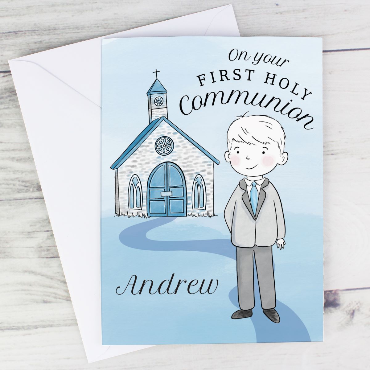 A personalized first holy communion greeting card and matching envelope on a light wood surface. The card features a cartoon illustration of a young boy in a suit standing in front of a church, with the text 'On your FIRST HOLY Communion' and the name 'Andrew'.