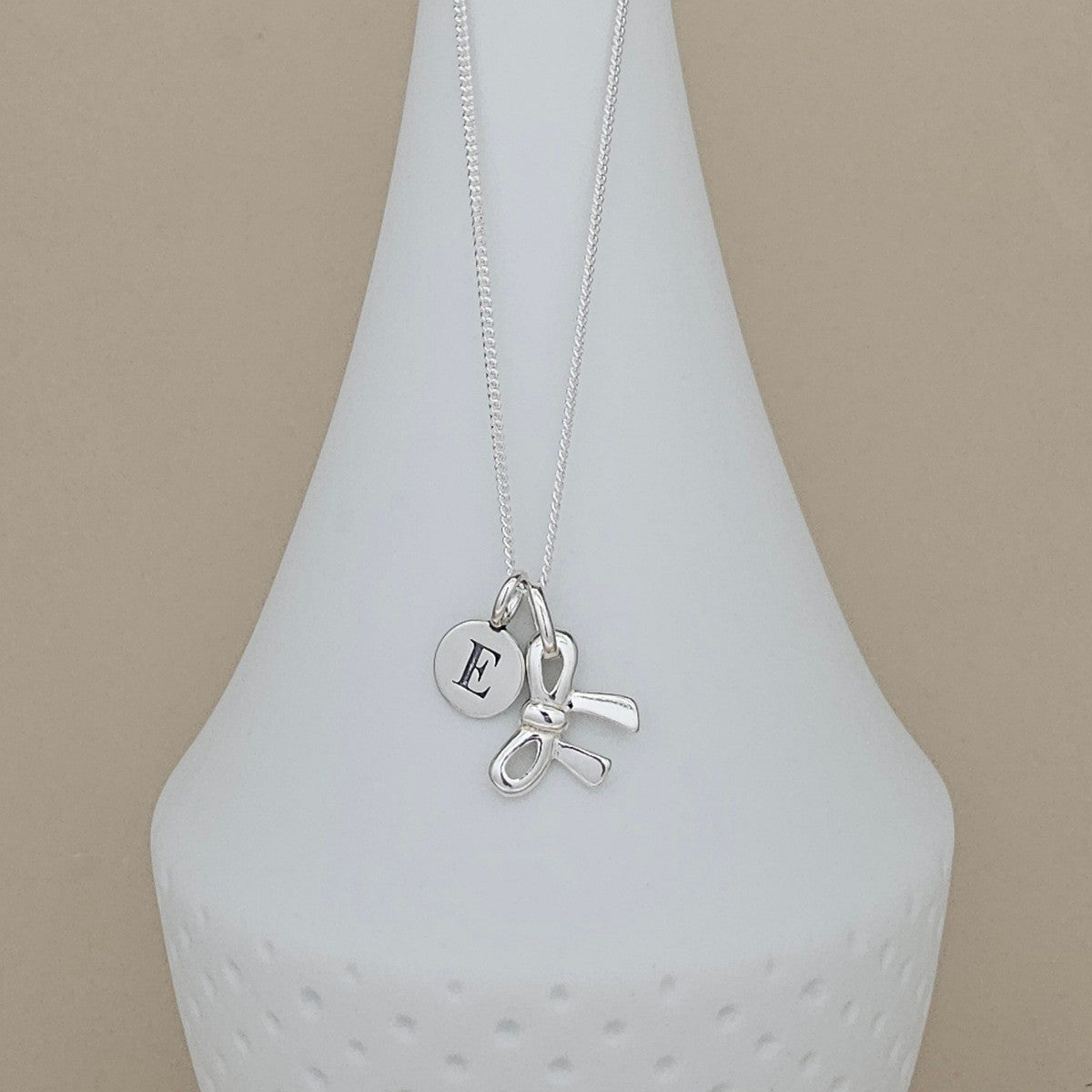 A silver necklace with a delicate chain, featuring a small round engraved silver disc with the letter 'E' and a bow-shaped charm, displayed on a white ceramic vase against a beige background.