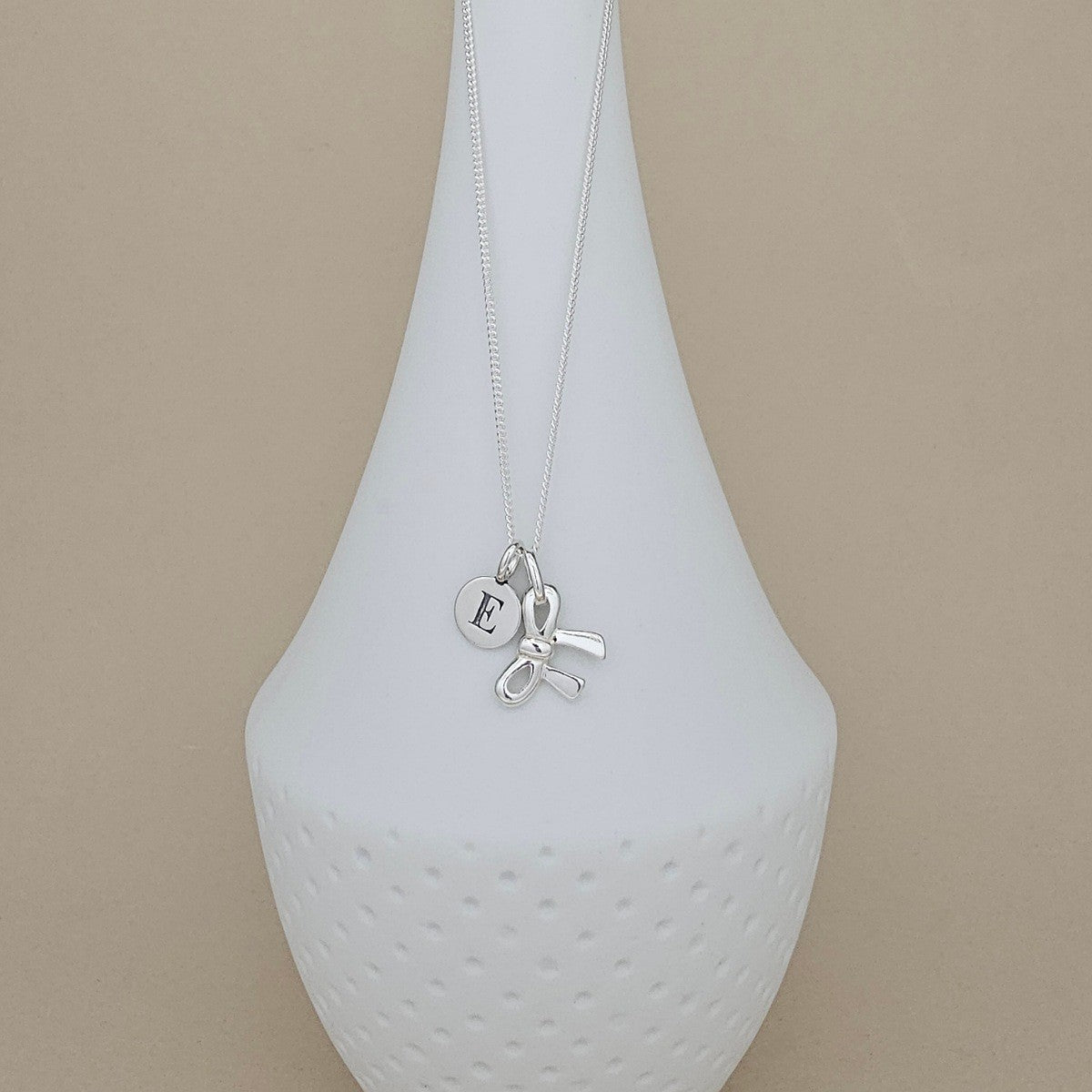 A silver necklace with a delicate chain and a charm featuring a small round initial 'E' pendant and a bow-shaped charm, displayed on a white ceramic vase against a beige background.
