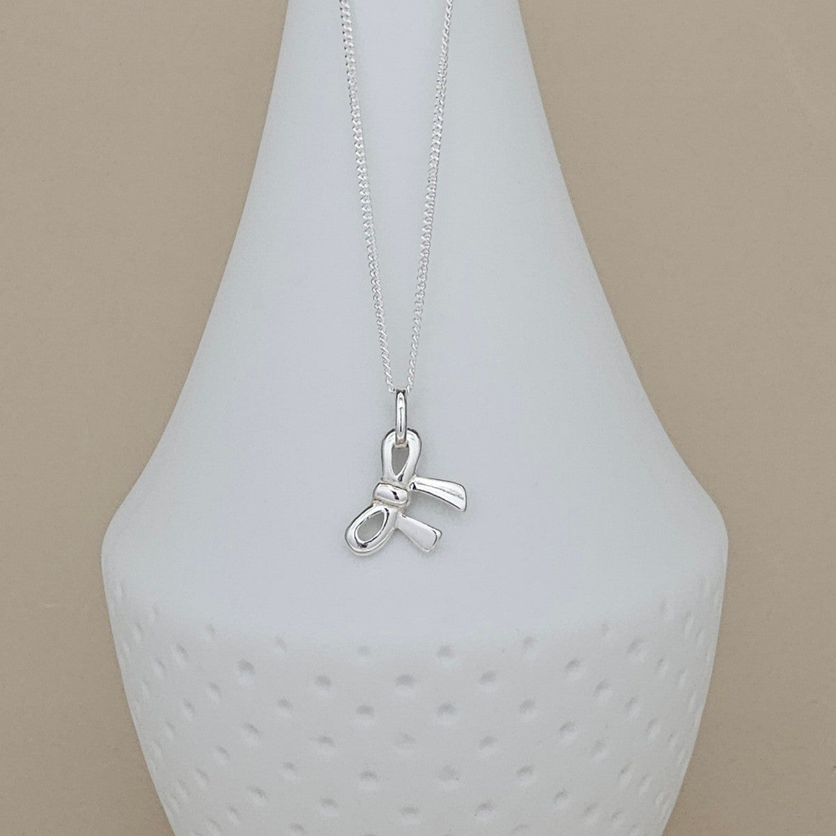 A silver necklace with a small bow-shaped pendant displayed on a white textured vase against a beige background.
