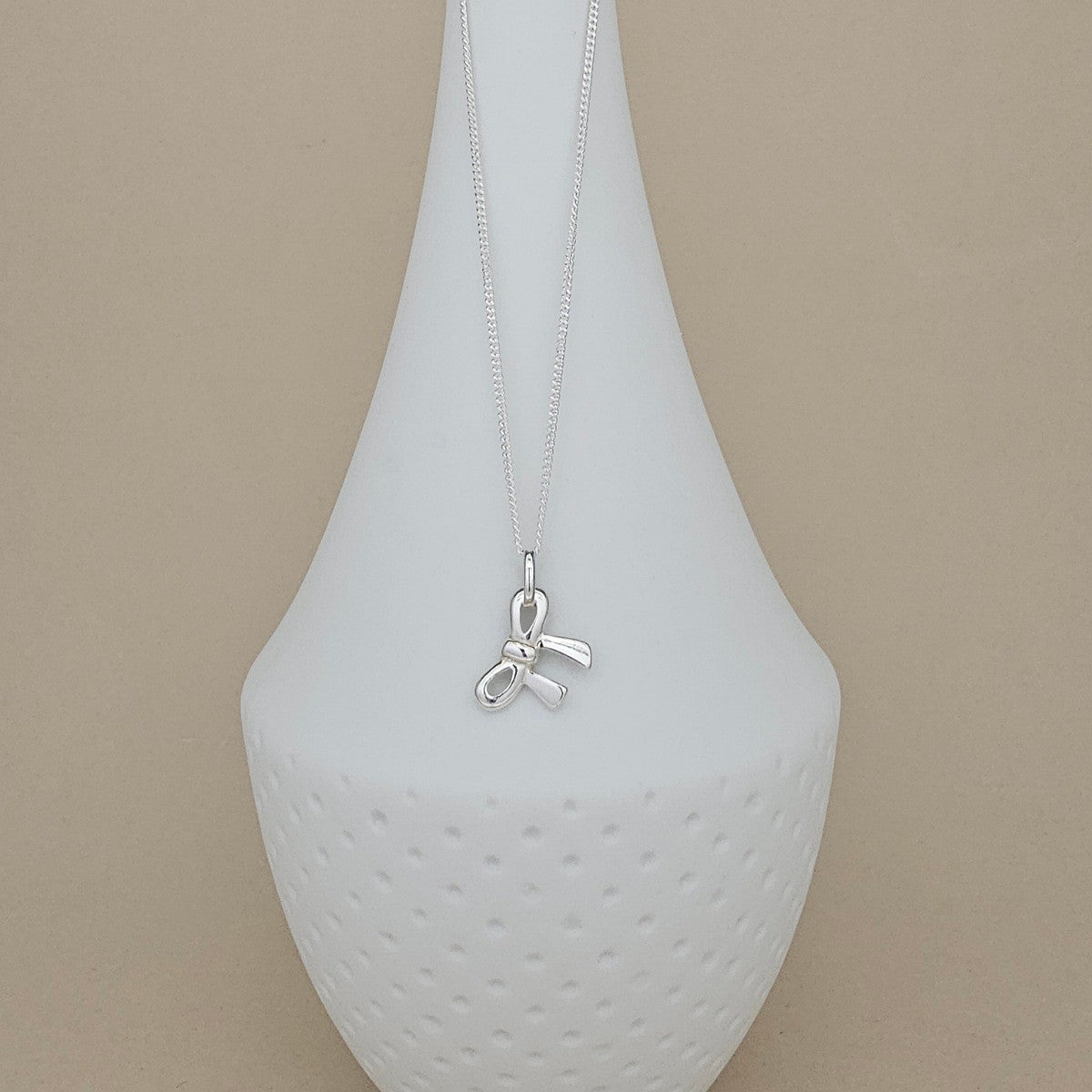 A silver bow pendant necklace displayed on a white ceramic vase against a warm beige background.