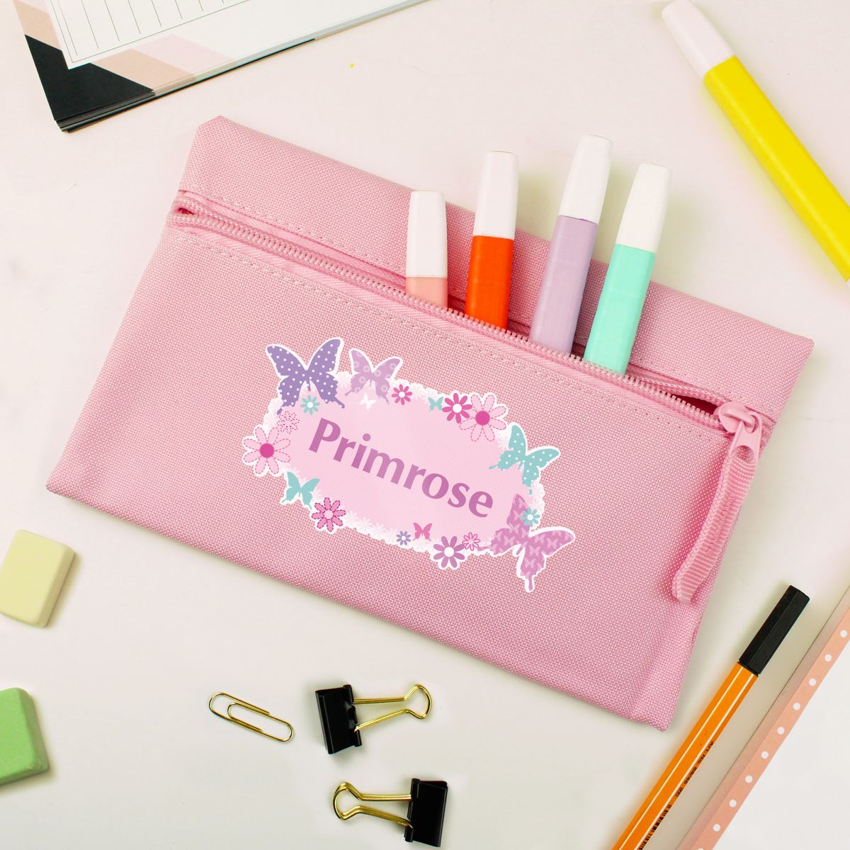A pink zippered pencil pouch with a butterfly and floral design on the front, personalized with the name 'Primrose'. It is shown open, containing four colored pens, surrounded by school supplies on a white desk.