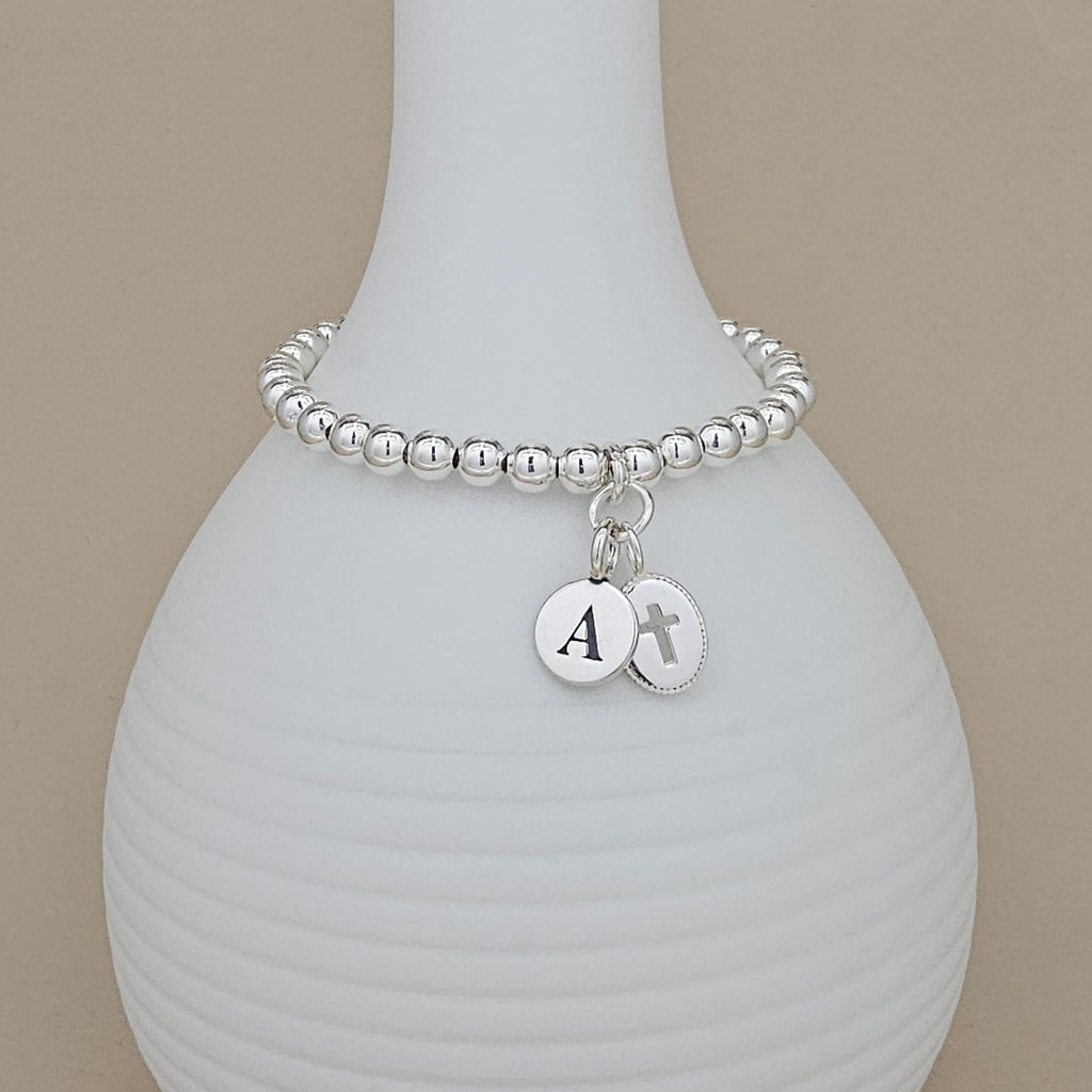 A silver beaded bracelet with two round silver charms, one engraved with the letter 'A' and the other with a cross, displayed on a white ribbed vase against a beige background.