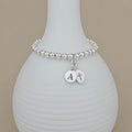 A silver beaded bracelet with two round silver charms, one engraved with the letter 'A' and the other with a cross, displayed on a white ribbed vase against a beige background.