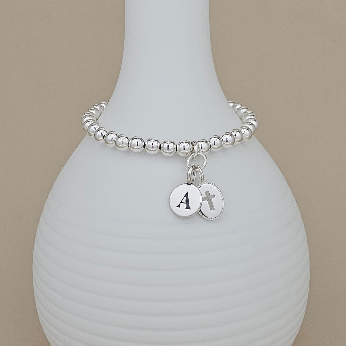 A silver beaded bracelet with two round silver charms, one engraved with the letter 'A' and the other with a cross, displayed on a white ribbed vase against a beige background.