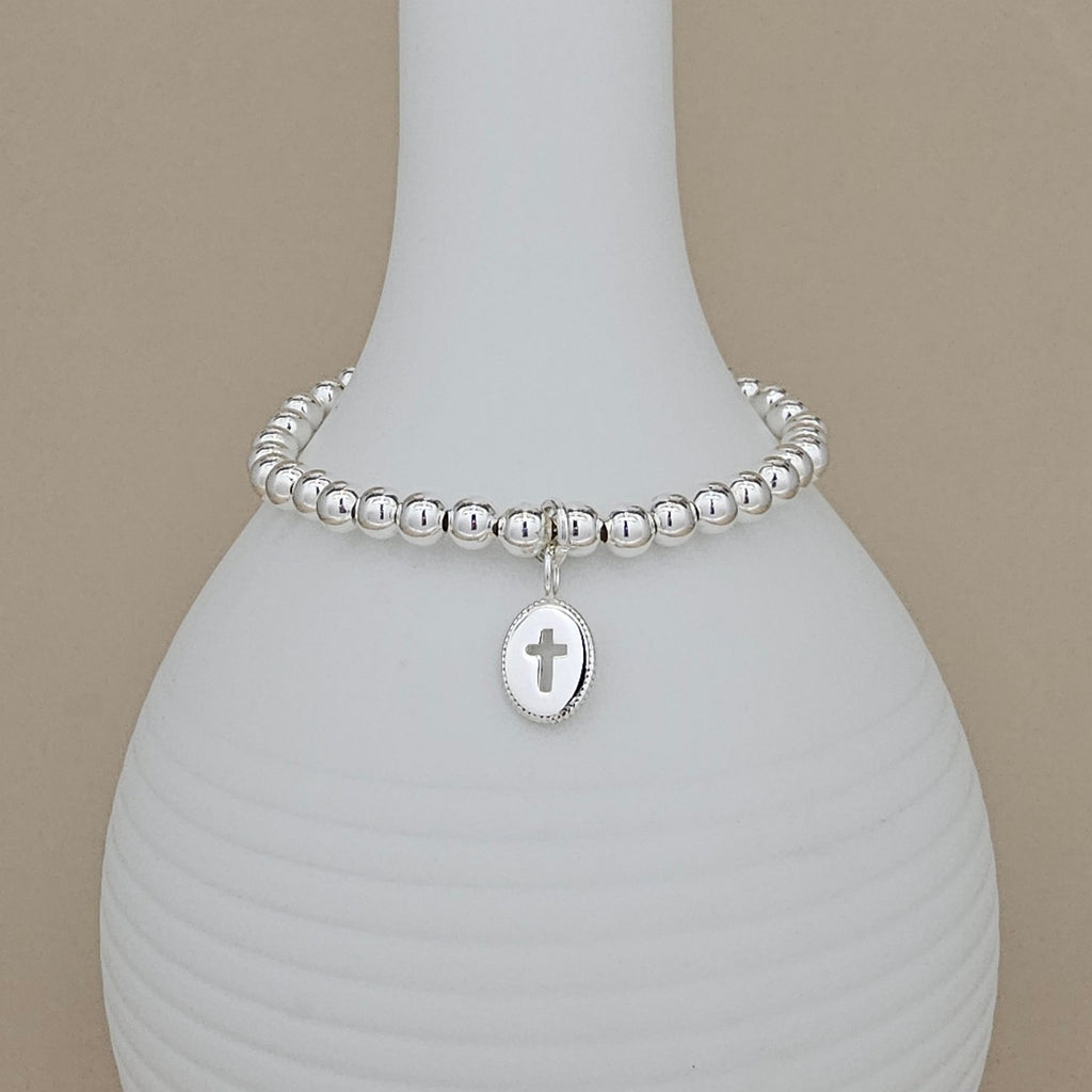 A silver beaded bracelet with a small oval cross pendant, displayed on a white ribbed vase against a beige background.