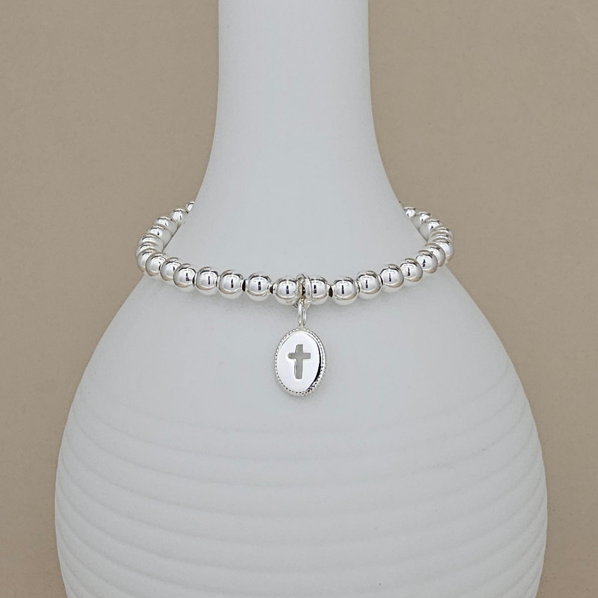 A silver beaded bracelet with a small oval cross pendant, displayed on a white ribbed vase against a beige background.